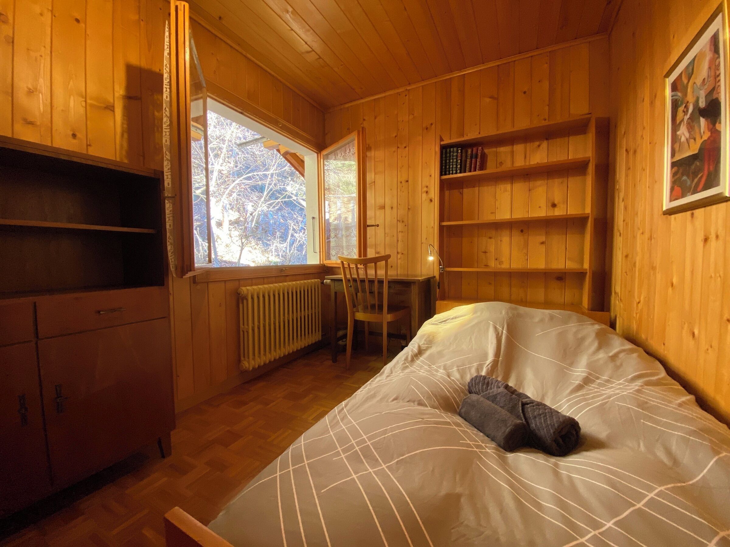 A wooden cabin room with a bed, a chair, a desk, and bookshelves.