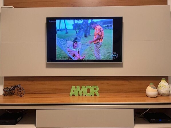 Un téléviseur fixé au mur, avec une étagère en bois en dessous sur laquelle est inscrit le mot « AMOR ».