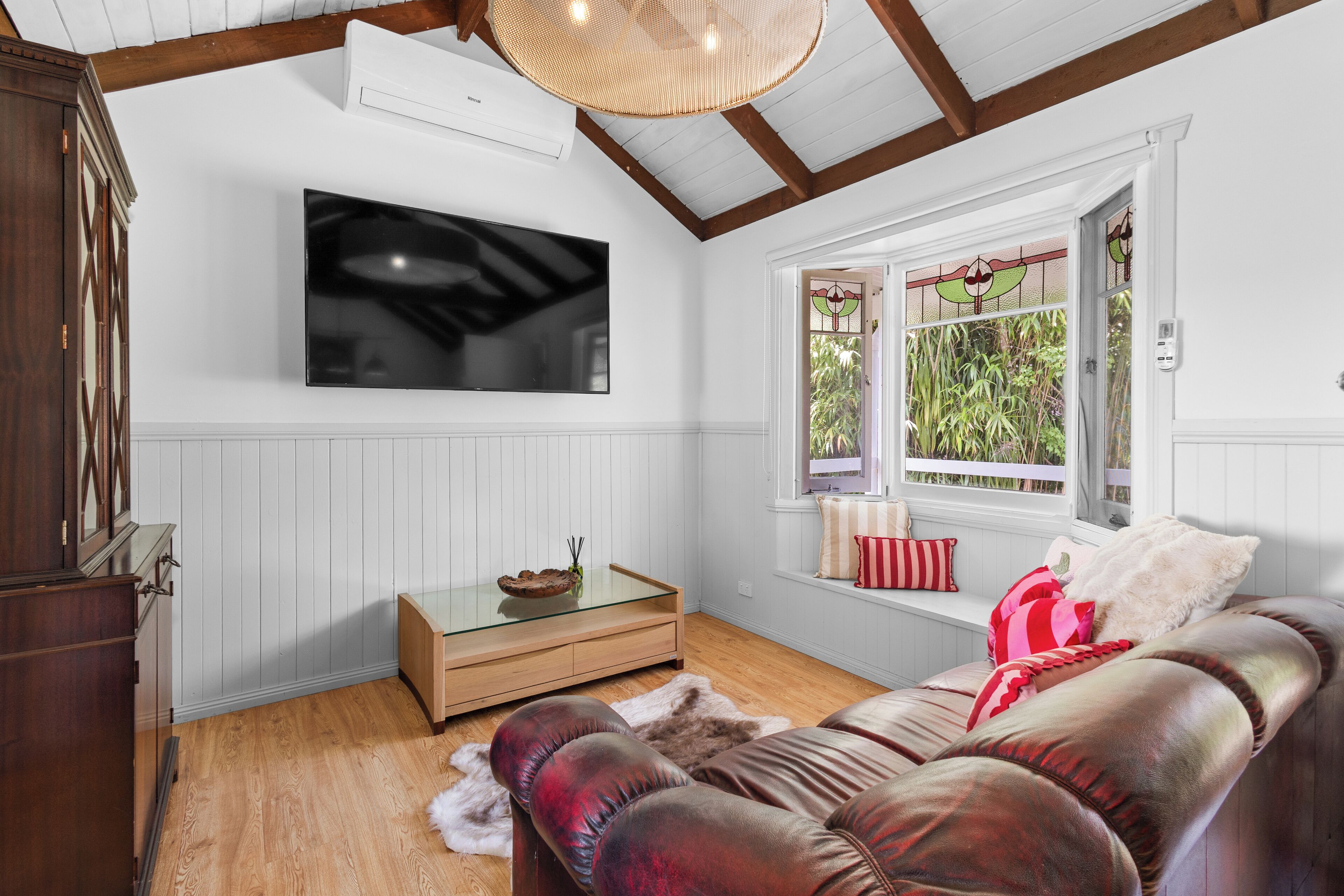 A living room with a flat-screen TV, wooden furniture, a glass coffee table, and a window with a view of greenery.