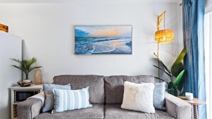 En stue med en grå sofa, et maleri af en strand, et lille bord med en plante og en lampe.