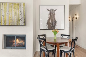 A dining area with a round table, four chairs, a vase with flowers, a fireplace, and two framed artworks on the wall. - Top-Floor Aspen Condo One Block from Gondola with Fireplace & Vaulted Ceilings (Aspen)