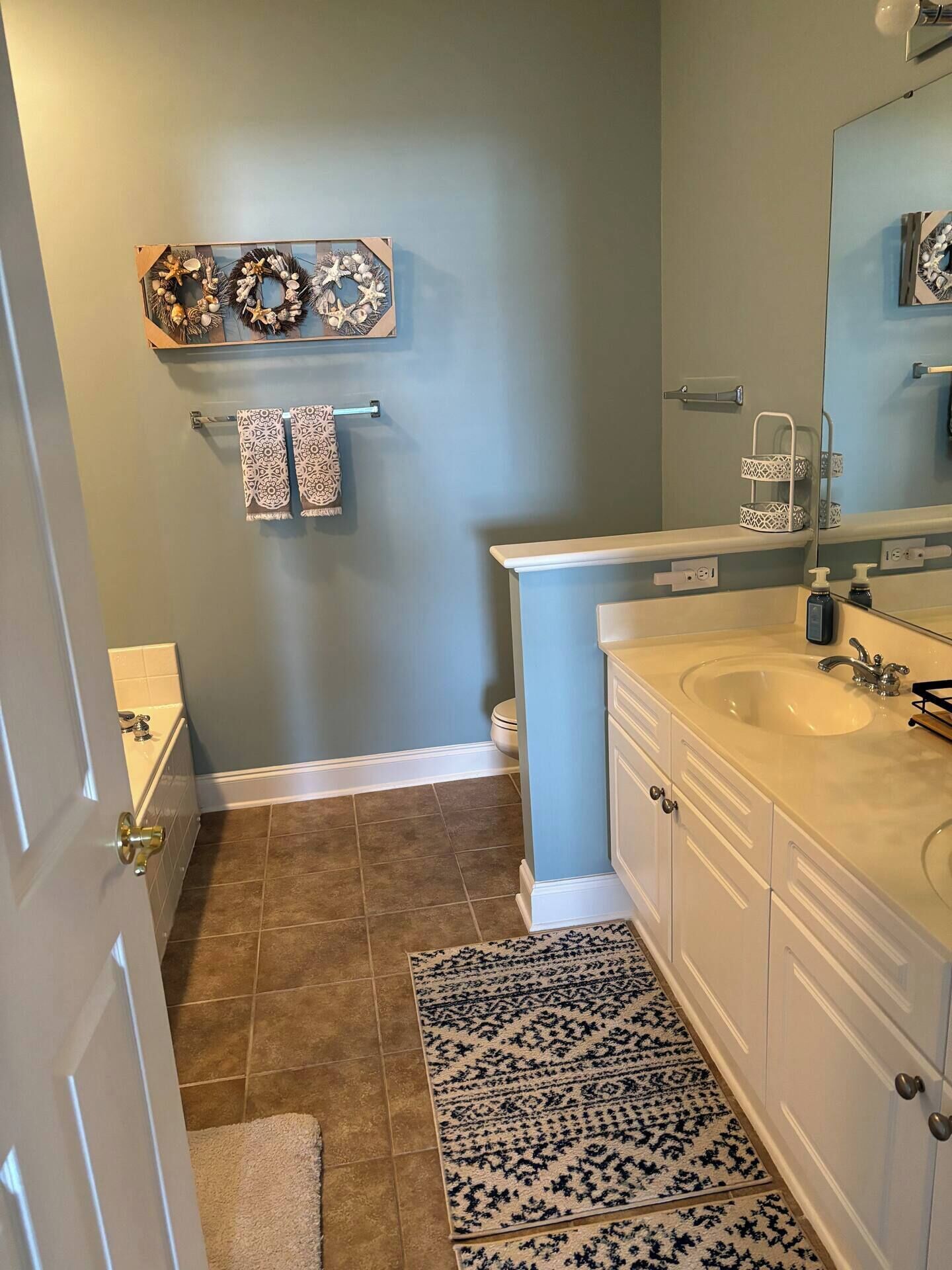 A bathroom with a double sink, a towel rack with patterned towels, a decorative wall piece above the sink, and a small rug on the floor.