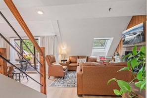 A modern living room with a sofa set, a dining area with chairs, a staircase, and a large window. - Fully Renovated Upscale Condo, Walk to Snowshed, Amenities Downstairs (Killington)