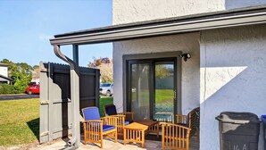 A covered patio with wooden furniture, a trash bin, and a sliding glass door leading to a grassy area.
