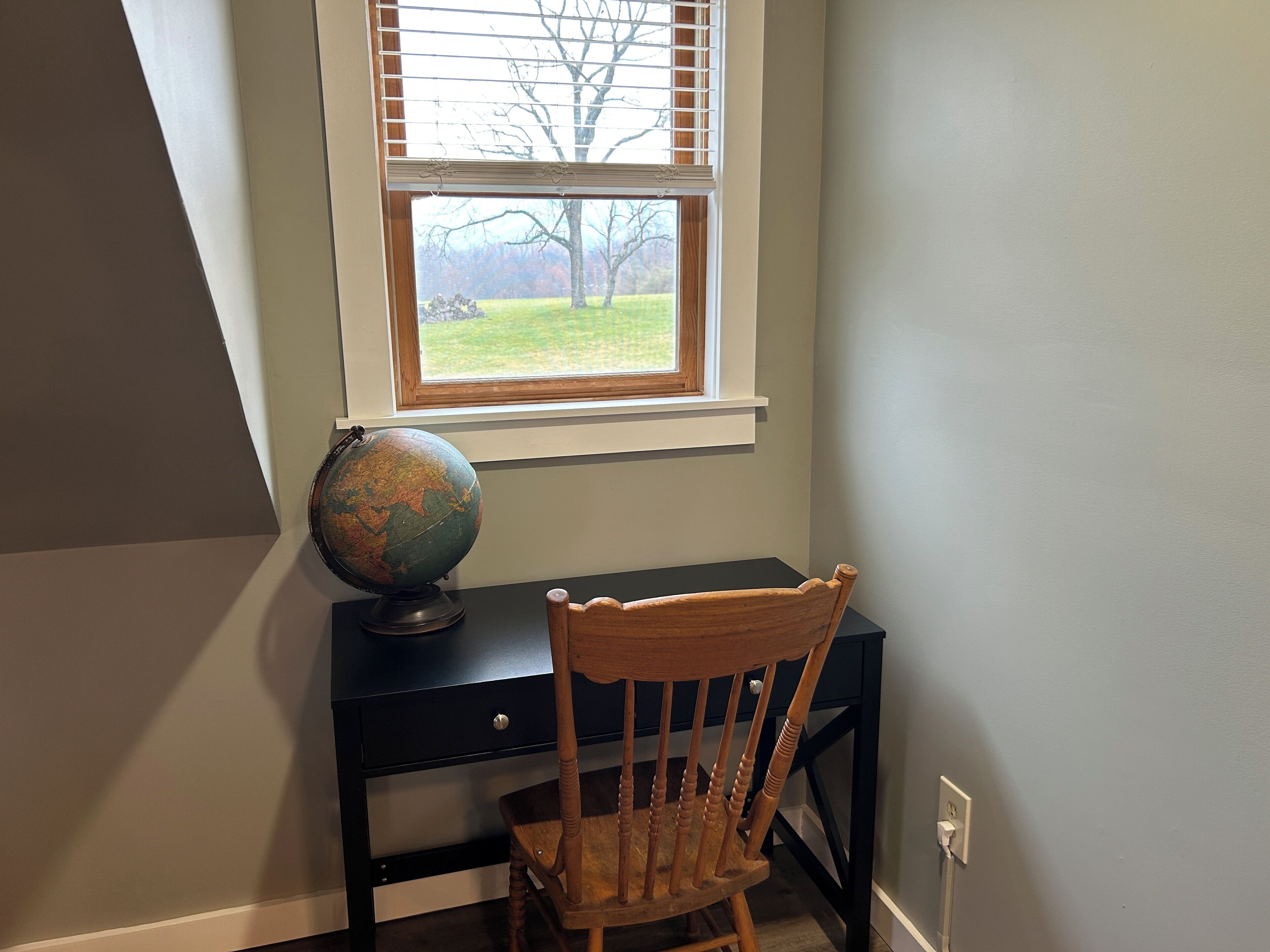 A desk with a globe and a wooden chair, positioned under a window with blinds, overlooking a grassy area.