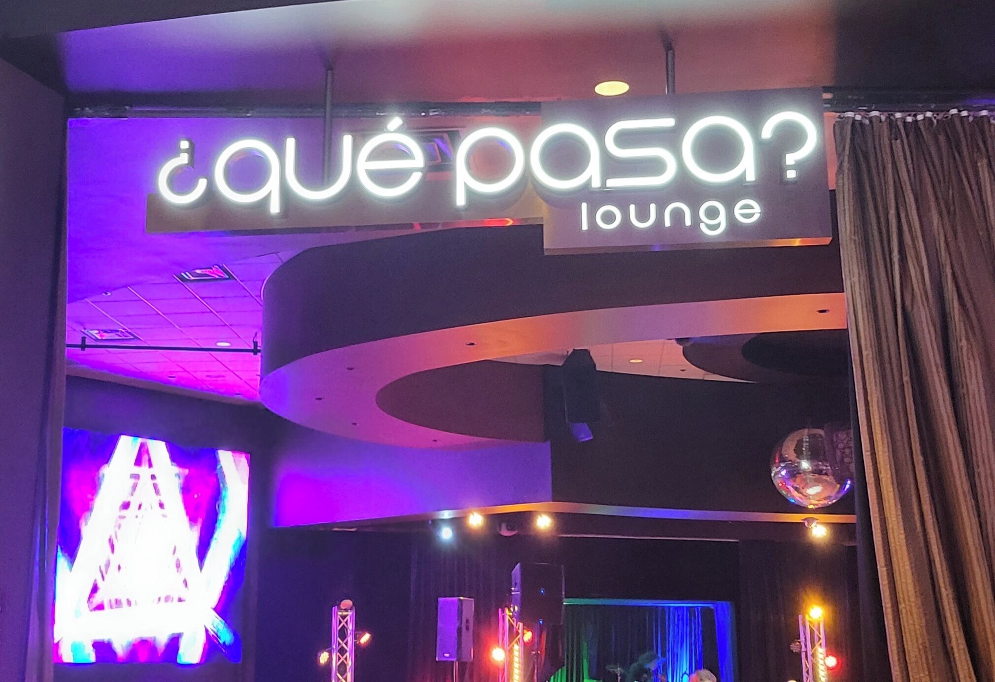 Oleskelualue, jossa on neonvalomainos, jossa lukee „¿qué pasa? lounge”, sekä lava valaistuslaitteineen.