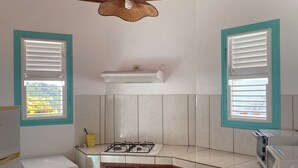 Une cuisine avec un plafond en bois, des armoires blanches, une cuisinière à gaz, un four à micro-ondes et deux fenêtres équipées de stores.