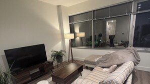 Una sala moderna con un televisor de pantalla plana grande, una mesa de centro de madera oscura, dos otomanas blancas, un sofá a rayas y una lámpara de pie.