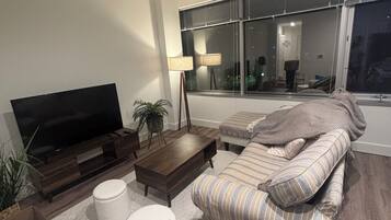 A modern living room with a large flat-screen TV, a dark wooden coffee table, two white ottomans, a striped sofa, and a floor lamp.