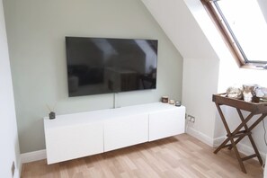 A modern living room with a large flat-screen TV mounted on a white wall, a wooden floor, and a skylight. - The Lily Suite in Berkshire (Wokingham)