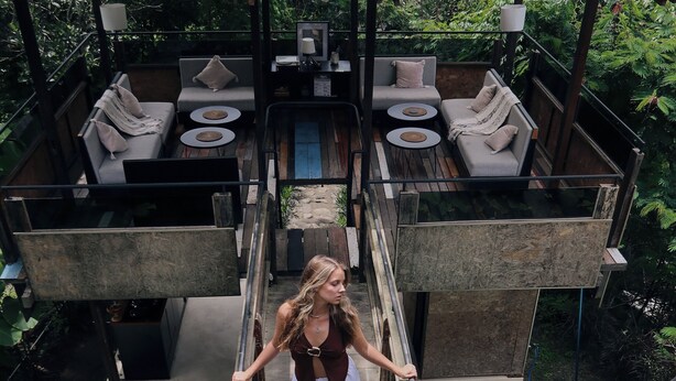 Eine Frau steigt eine Holztreppe in einem Resort hinunter, das Außenbereichs-Sitzgelegenheiten und einen Pool bietet.