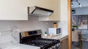 Une cuisine moderne équipée d’une cuisinière à gaz, d’un four et de meubles blancs. On aperçoit également un micro-ondes, une petite plante et un coin repas à l’arrière-plan.