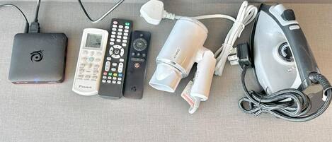 Ein Flachbildfernseher, ein mobiler Heizlüfter, ein Staubsauger, eine Fernbedienung, eine Mehrfachsteckdose und eine Kabelbox auf einer Stoffoberfläche.