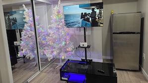 A modern room with a white Christmas tree, a flat-screen TV, a refrigerator, and a glass table with blue lighting.
