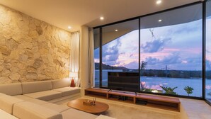 A modern living room with a stone wall, a large window with a view of a body of water, and a flat-screen TV on a wooden stand. - Villa Kalani Soul by Alfred in Bali (Pecatu)