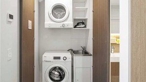 A compact laundry room with a front-loading washer and dryer, a small sink, and built-in shelving. - River View High-Rise 1BR Apartment by GC Casino (Broadbeach)
