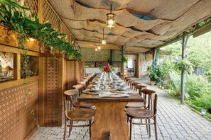 Ein langer Holztisch ist für das Essen gedeckt, mit Stühlen versehen, umgeben von einer rustikalen Holzdecke und -wänden, mit Grünpflanzen und Blick ins Freie.
