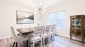 Un'area pranzo con un tavolo di legno e sedie bianche, un quadro appeso al muro, un lampadario e una finestra con vista.