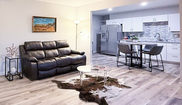 A modern living room with a leather sofa, a glass coffee table, and a cowhide rug. It leads to a kitchen with white cabinets and a bar area. - Lakefront + Hot Tub + Dock + Backyard Trail Access + 2 Min to OZ Bike Park (Bella Vista)