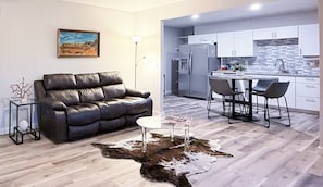 A modern living room with a leather sofa, a glass coffee table, and a cowhide rug. It leads to a kitchen with white cabinets and a bar area.