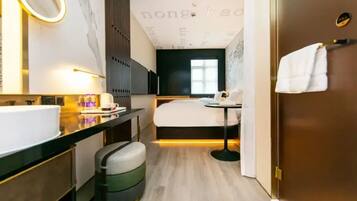 A modern hotel room with a large bed, a small table, a round mirror, a sink, and a trash bin.
