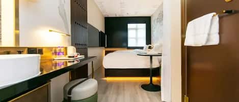 A modern hotel room with a large bed, a small table, a round mirror, a sink, and a trash bin.