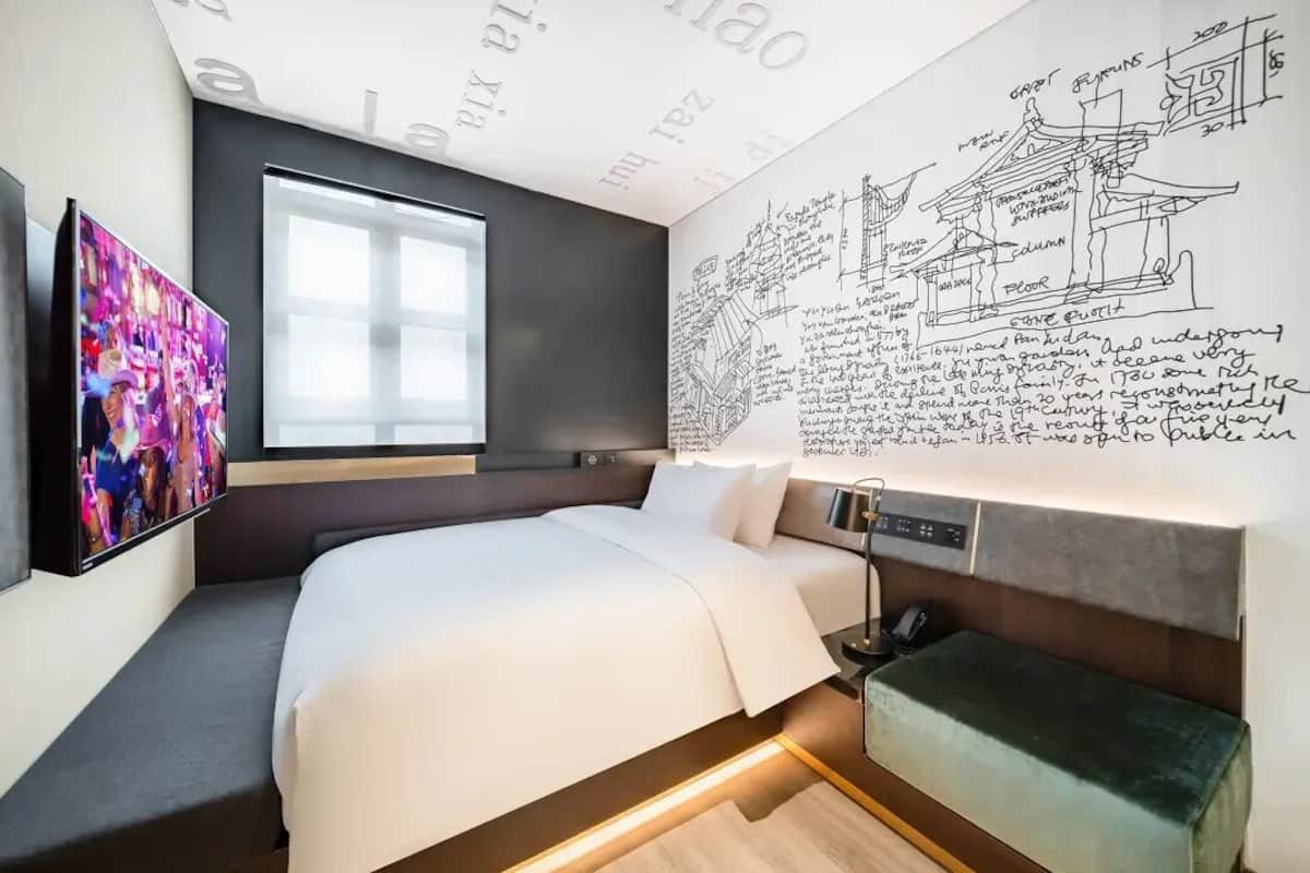Ein modernes Hotelzimmer mit einem großen Bett, einem Flachbildfernseher, einem Fenster und einer Wand mit architektonischen Zeichnungen.