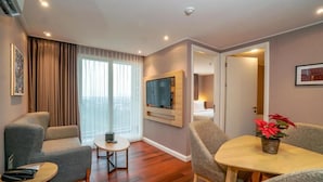 A modern living room with a flat-screen TV mounted on the wall, a grey sofa, a small round table, and a potted plant. - Swiss-Belcourt Serpong (South Tangerang)