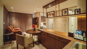 A modern kitchen with wooden cabinets, a dining area with a table and chairs, and a living area with a sofa and a TV. - Swiss-Belcourt Serpong (South Tangerang)