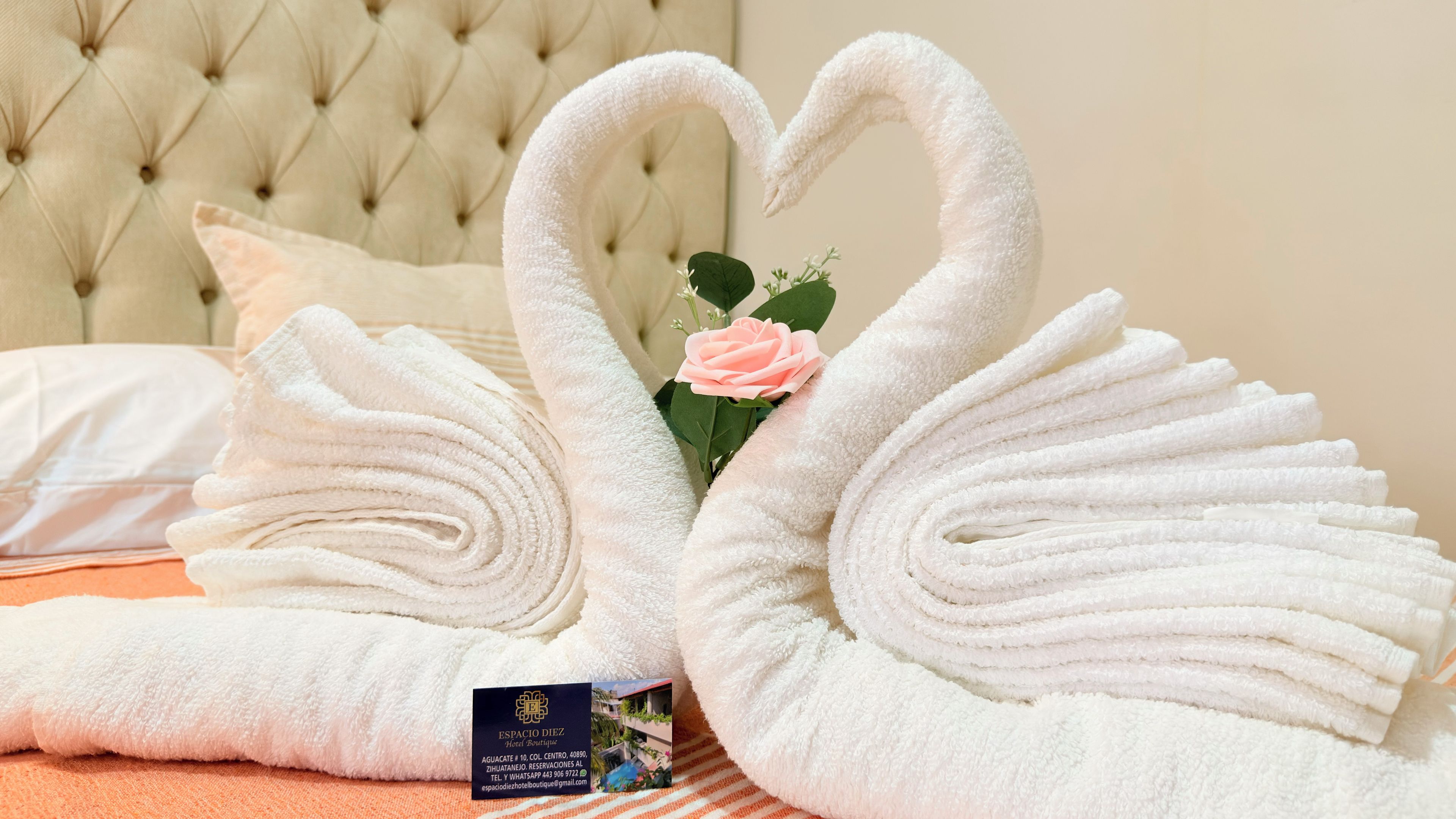 A hotel room with a neatly folded white towel arranged in the shape of a swan, a pink rose, and a business card on the bed.
