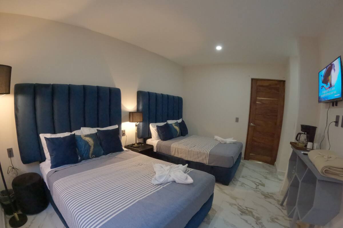 A hotel room with two beds, each with a headboard and pillows, a small desk with a chair, a television mounted on the wall, and a door.