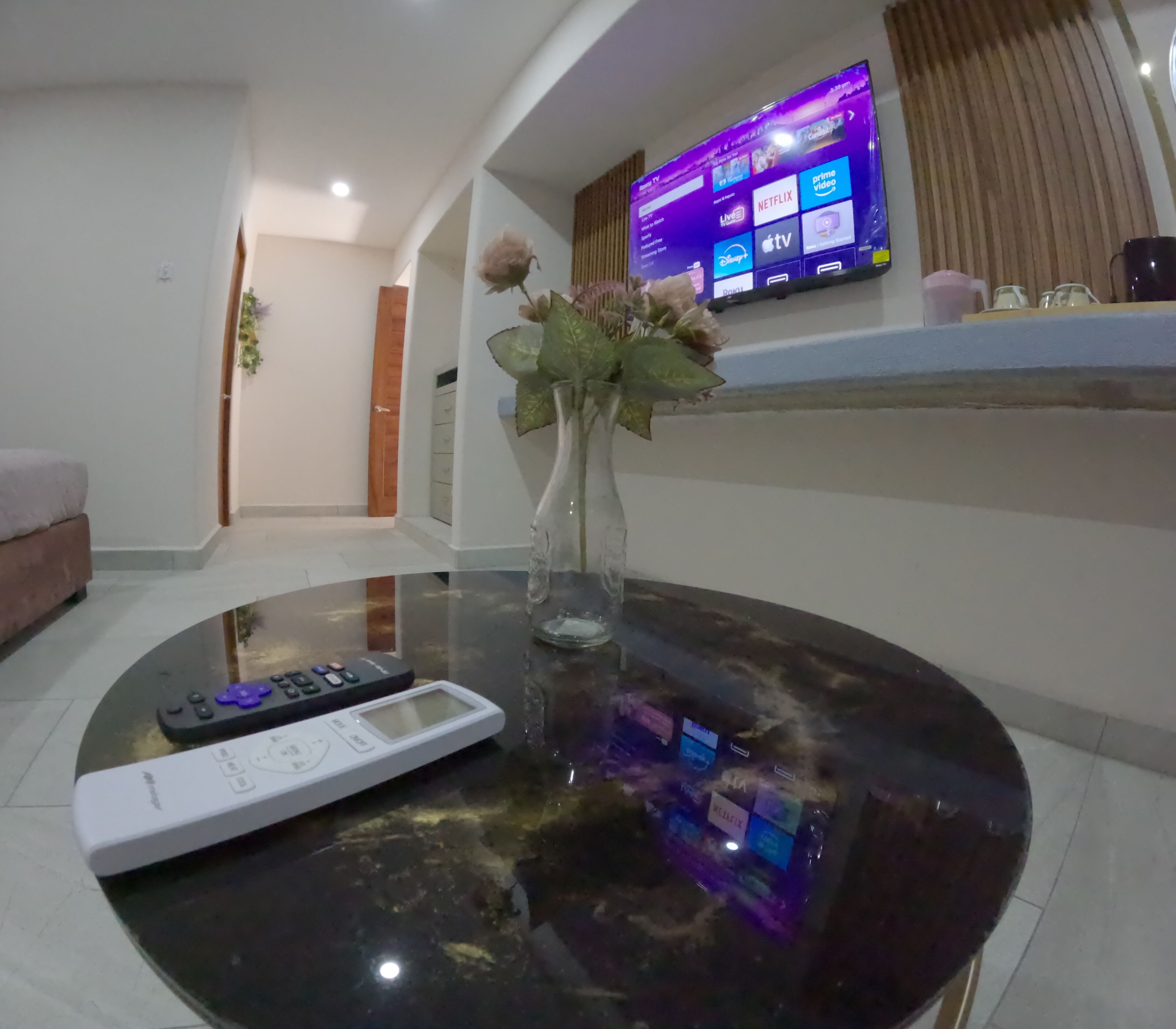 A living room with a round glass coffee table, a remote control, and a vase with flowers.