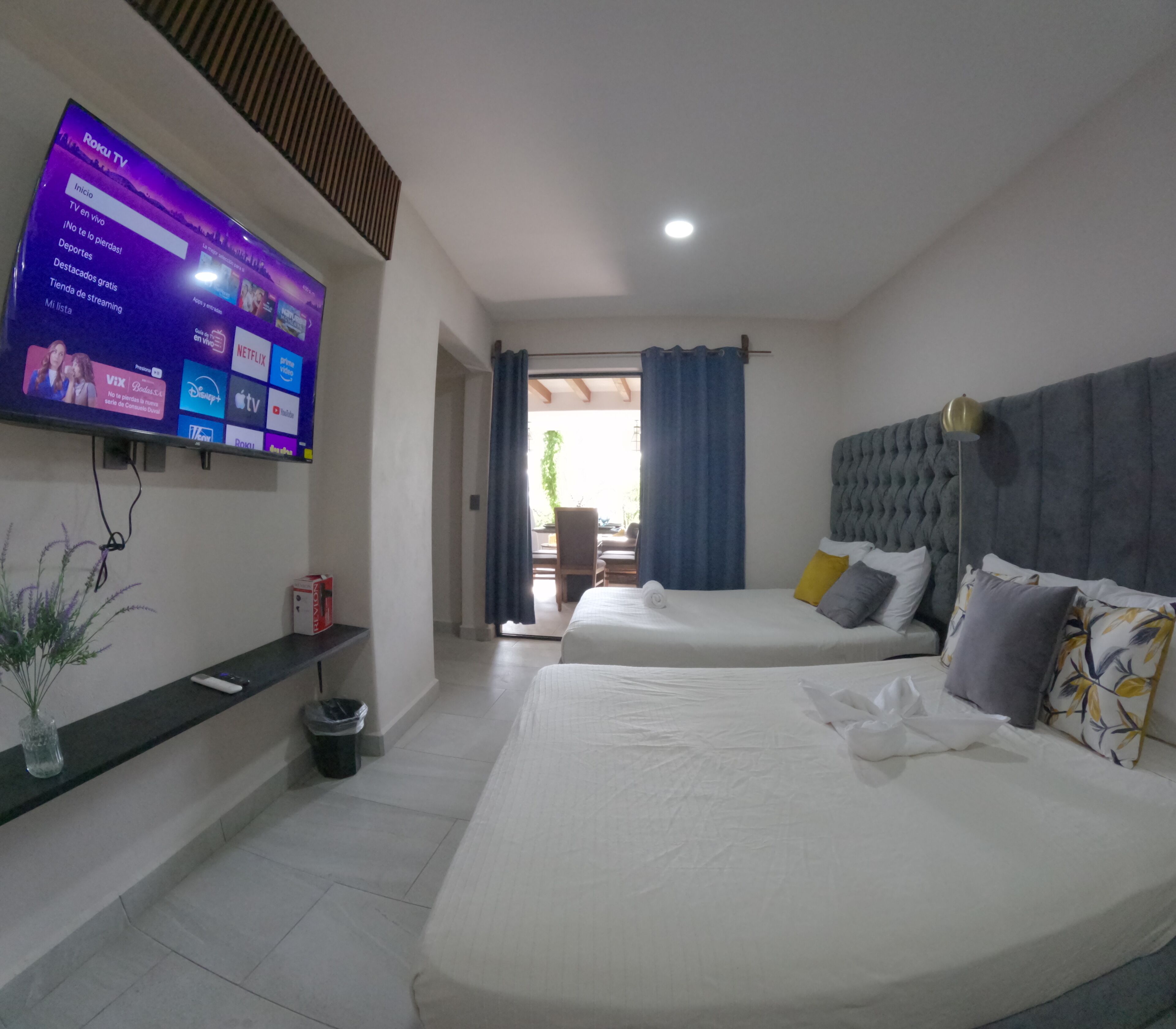 Quarto de hotel com duas camas, uma TV fixada na parede exibindo vários canais, e vista para o exterior através de uma porta de vidro.