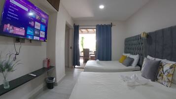 Quarto de hotel com duas camas, uma TV fixada na parede exibindo vários canais, e vista para o exterior através de uma porta de vidro.