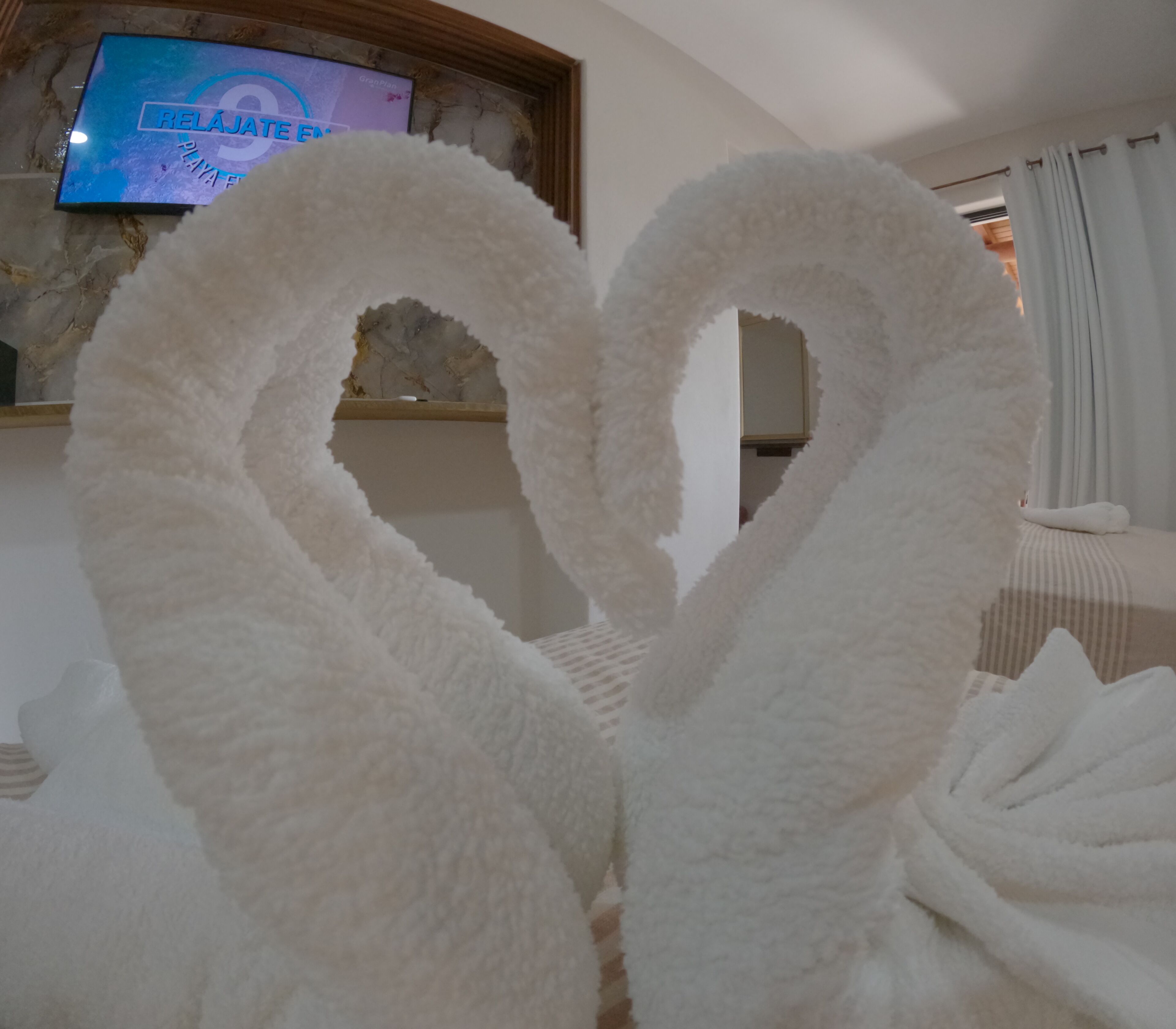 A room with a heart-shaped arrangement of white towels, a television displaying 'RELAJATE EN PLAYA', and a window with curtains.