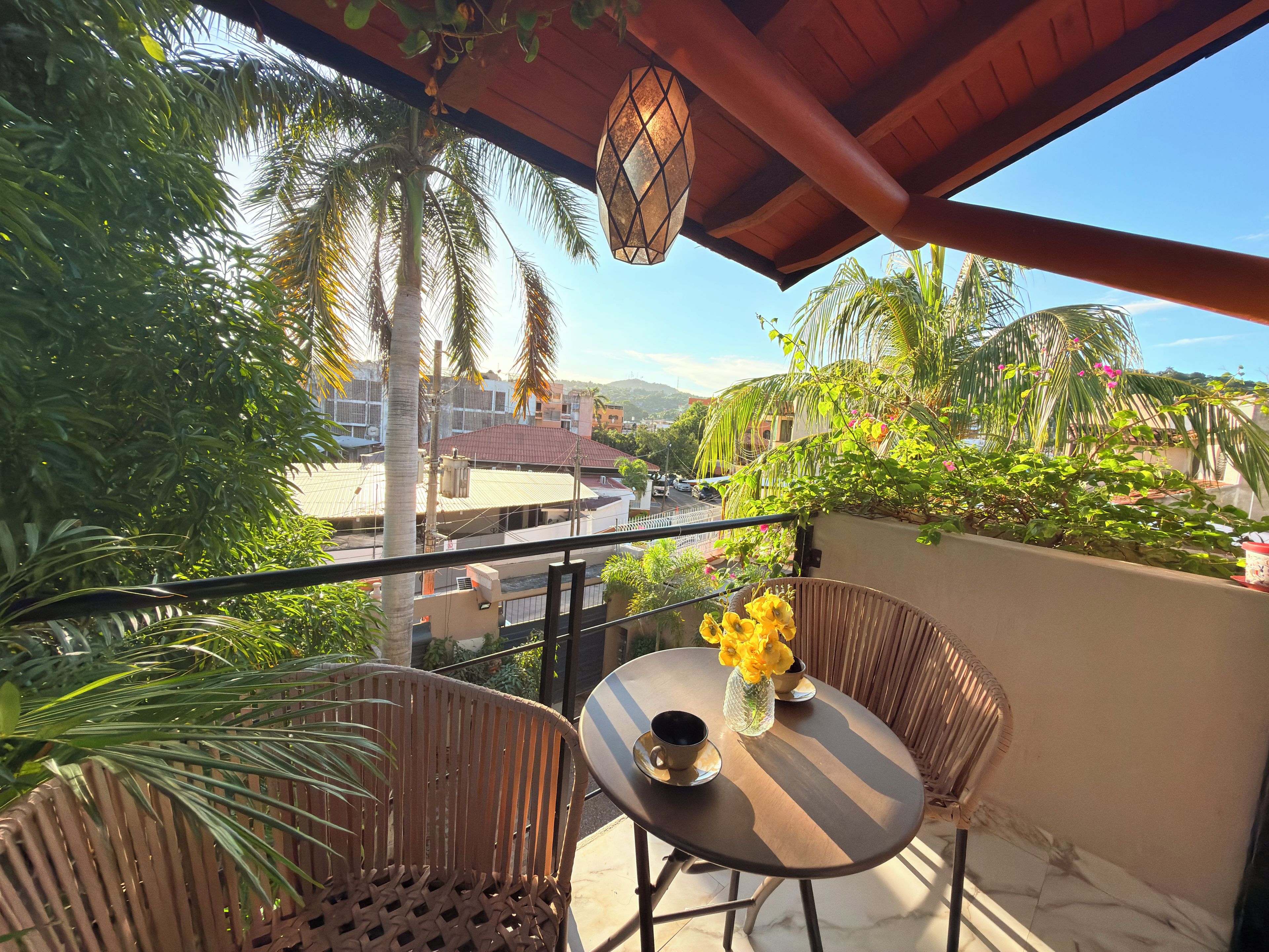 Un balcón con mobiliario de mimbre, una mesa con un florero y vistas a edificios y palmeras.