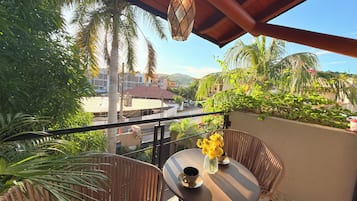 Un balcón con mobiliario de mimbre, una mesa con un florero y vistas a edificios y palmeras.