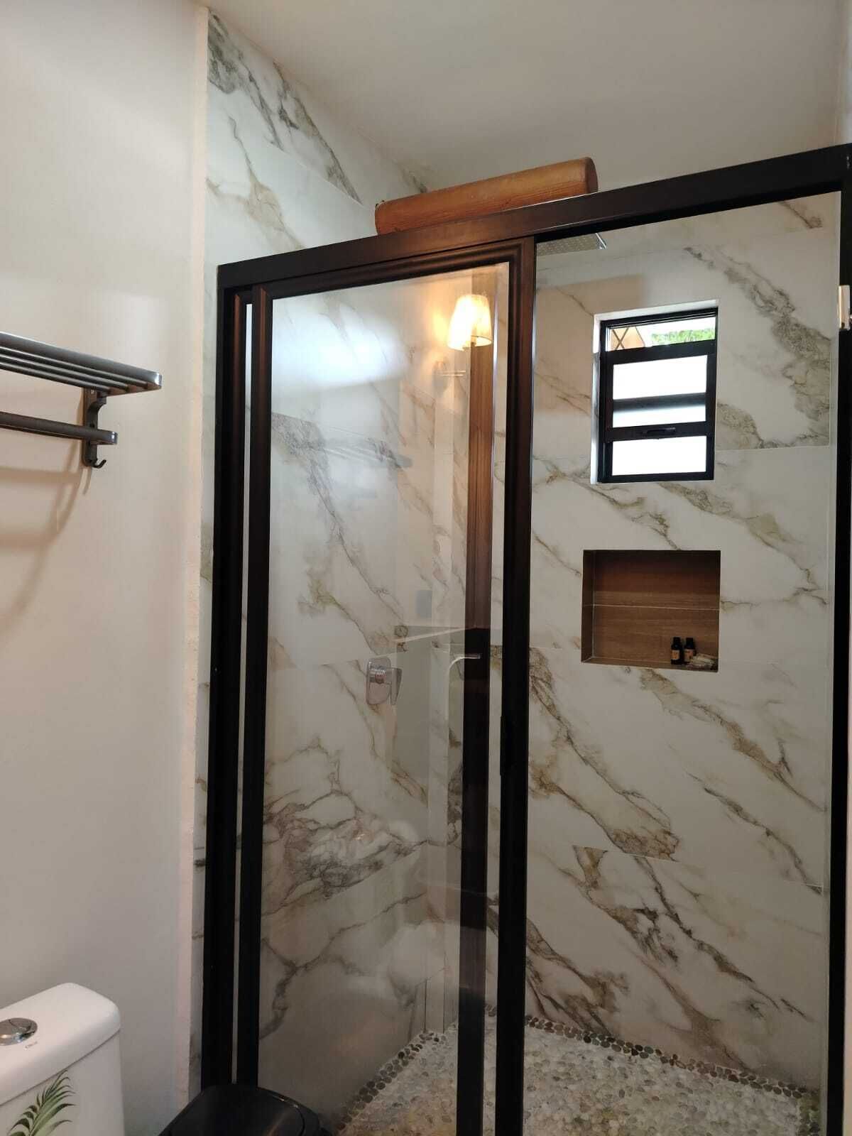 Un baño con ducha de mampara de vidrio, paredes de mármol, estante de madera y perchero.