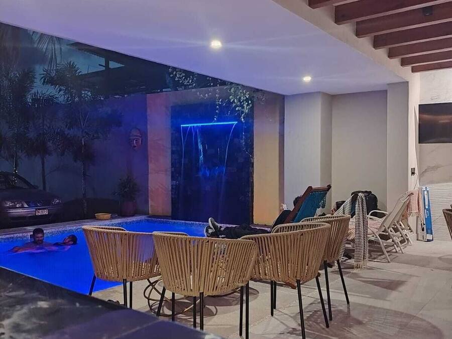 Área de piscina externa com cachoeira, cercada por cadeiras de vime e um bar.