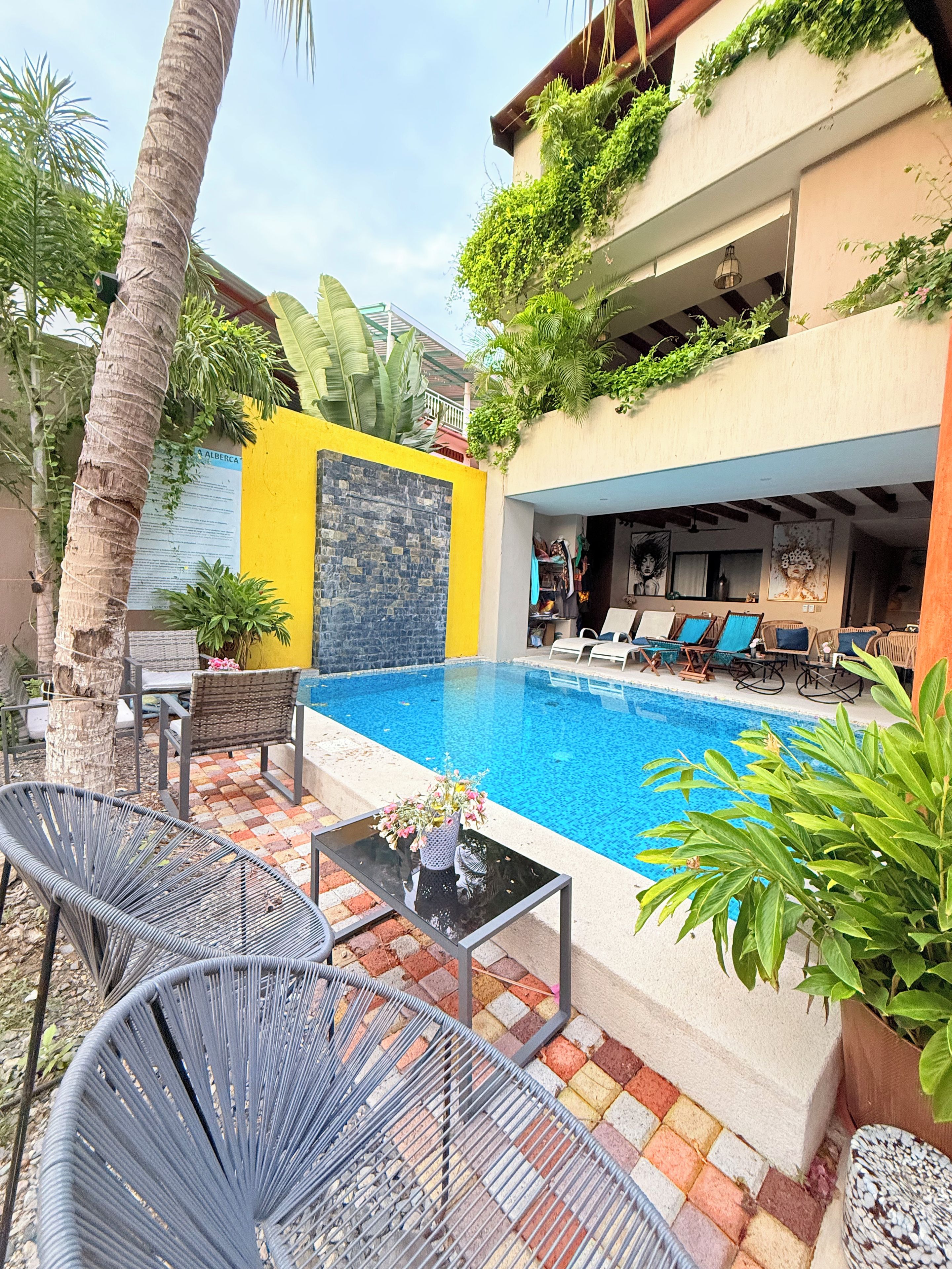 Un área de piscina con sillones de descanso y una mesa, rodeada de plantas y un muro amarillo.