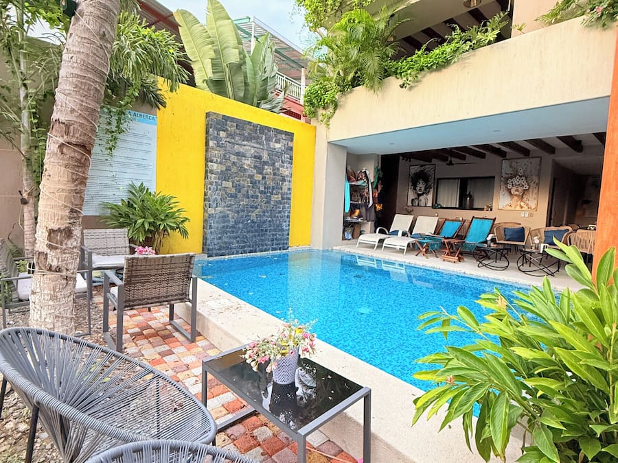 Un área de piscina con sillones de descanso y una mesa, rodeada de plantas y un muro amarillo.