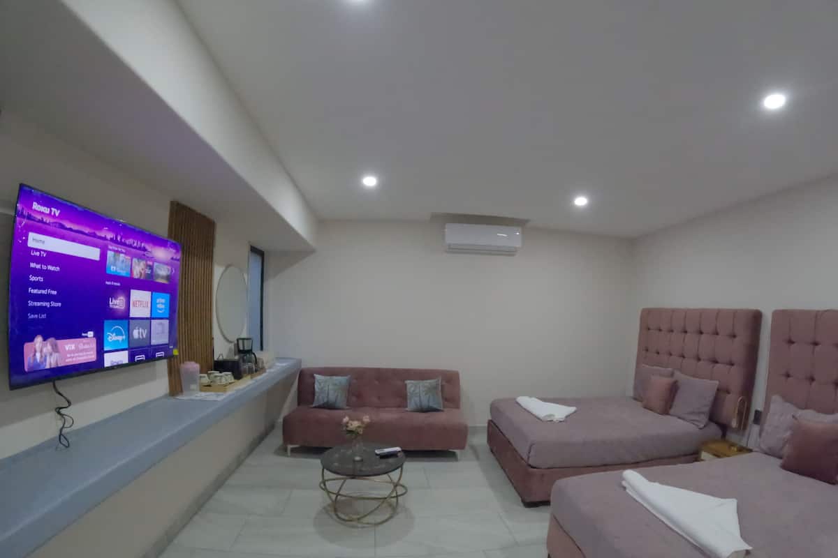 A modern hotel room with a flat-screen TV displaying a streaming service interface, a sofa, two beds, a small round table, and a ceiling-mounted air conditioner.