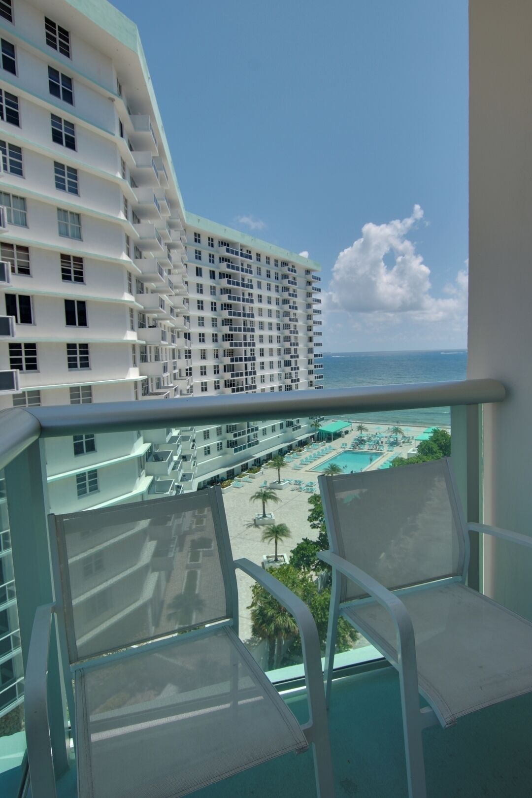 Een balkon met twee witte stoelen, met uitzicht op een zwembadgedeelte met palmbomen en een hoge wolkenkrabber.