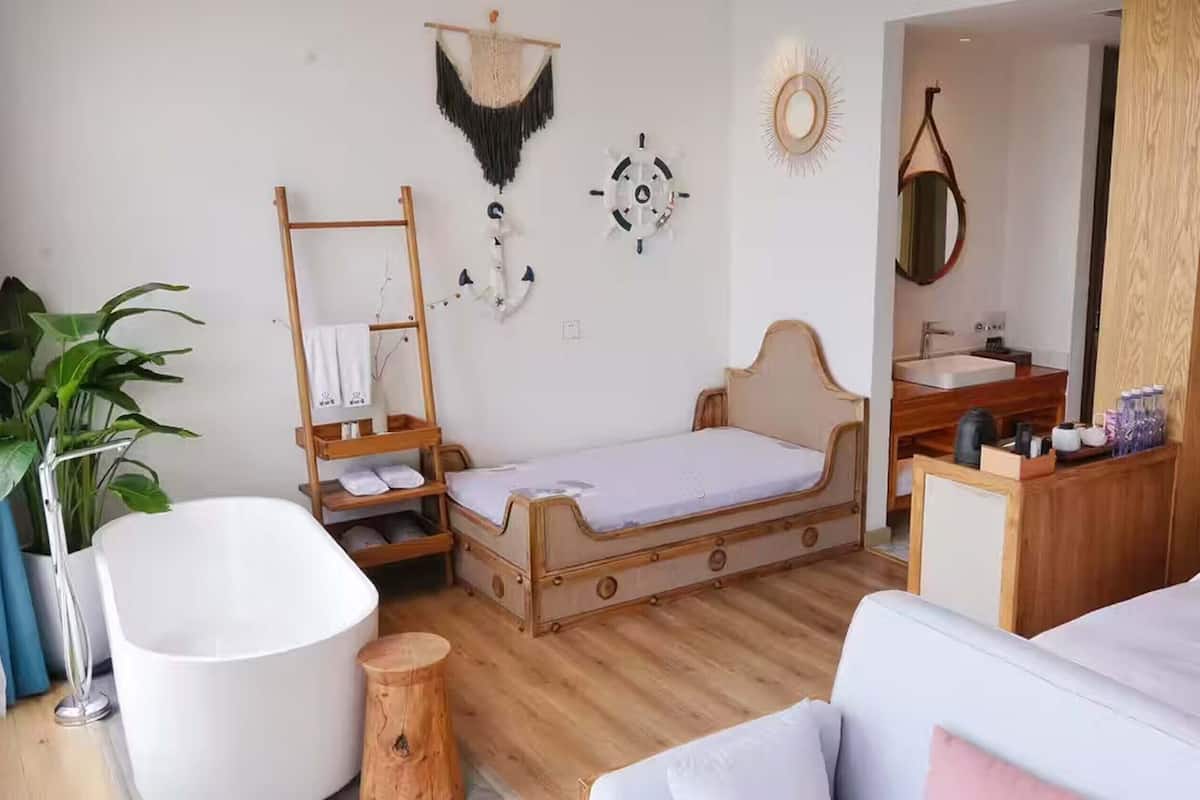 A hotel room with a wooden bed, a white bathtub, a wooden vanity with a mirror, and a large potted plant.