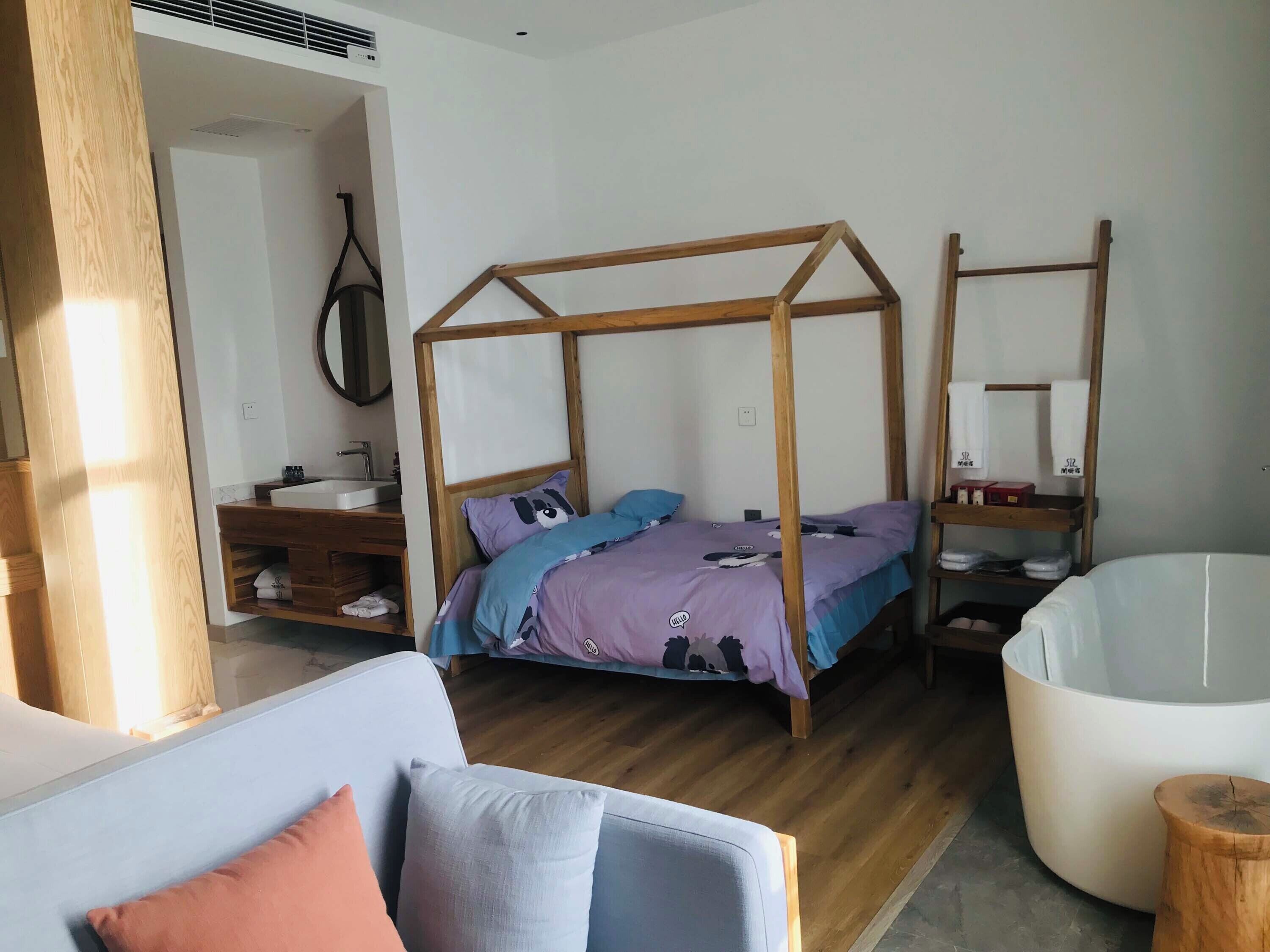 Een slaapkamer met een houten baldakijnbed, een wit ligbad, een houten kaptafel met spiegel en een witte bank met kussens.