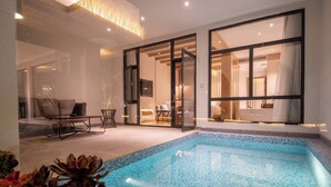 En moderne indendørs swimmingpool med klare blå fliser, et opholdsområde med sofa og stole, samt store glasdøre der fører til en stue med fjernsyn og spiseplads.