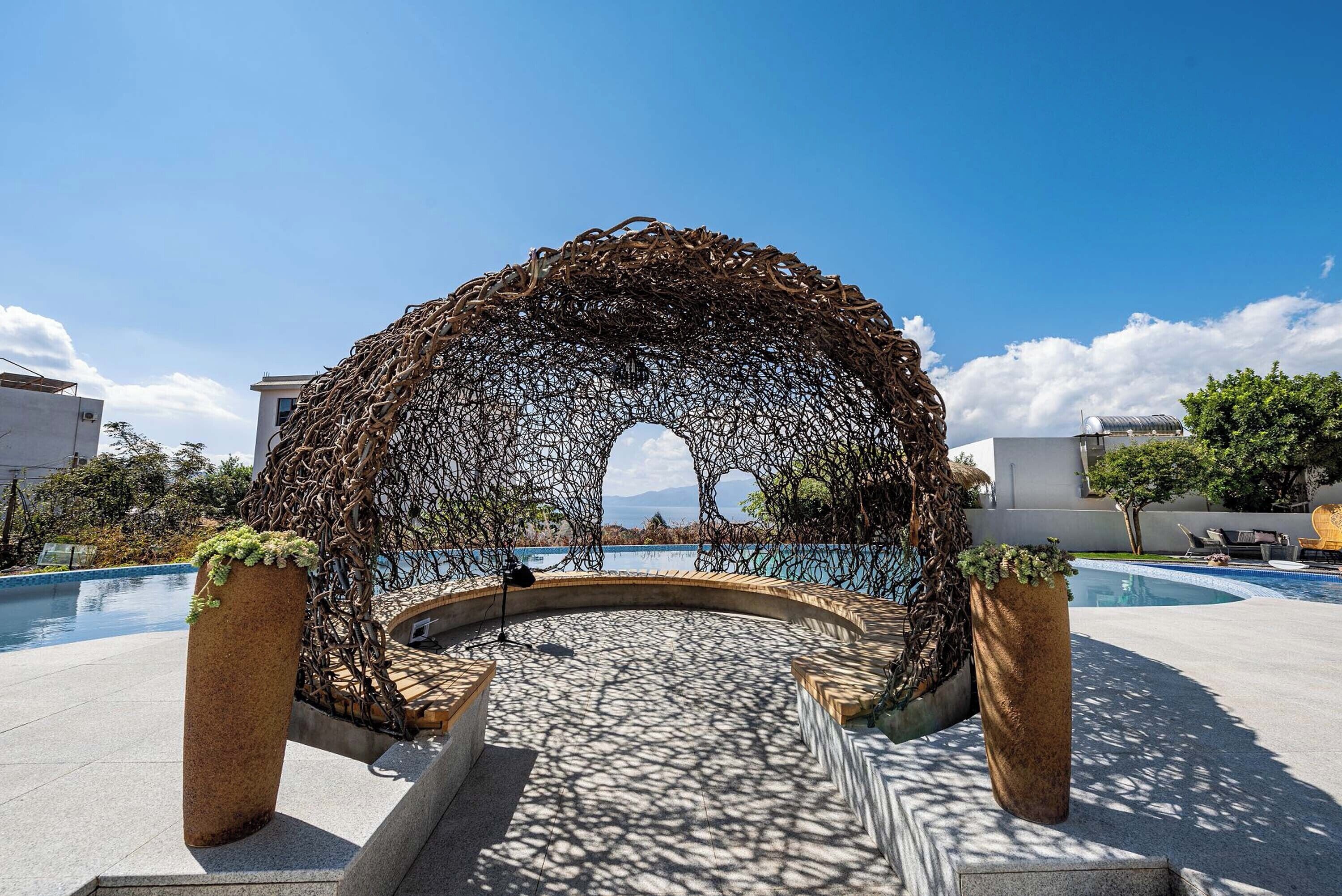 Un área de estar al aire libre con forma circular, con una estructura de arco de metal, rodeada por una piscina y plantas en macetas.