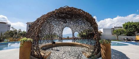 Un área de estar al aire libre con forma circular, con una estructura de arco de metal, rodeada por una piscina y plantas en macetas.