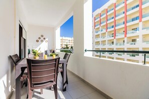 A balcony with wicker furniture, a dining table, and a view of a multi-story building. - Quinta das Palmeiras Pool & Beach Stay (Alporcinhos)