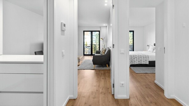 Un interior moderno con un pasillo que conduce a un dormitorio y una sala de estar, con piso de madera, paredes blancas y un ventanal grande.